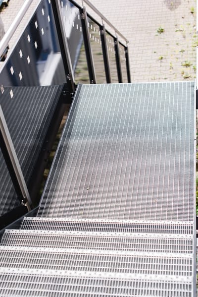 Detailaufnahme einer Außentreppe mit Gitterroststufen und Handlauf aus Metall.